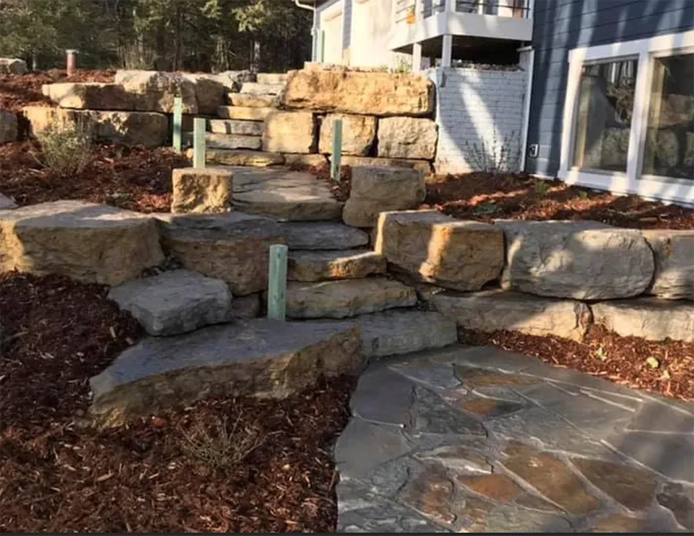 Stone steps leading up a mulched hill toward a house patio, with large boulders used as retaining walls.