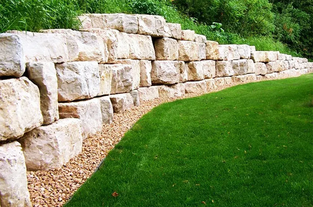 A stone retaining wall curves along the edge of a green lawn with a strip of gravel at its base.