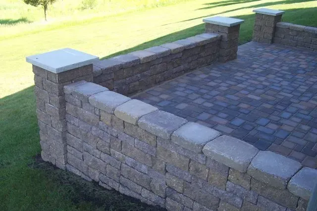 A patio constructed with multi-toned paver blocks featuring a retaining wall and corner pillars topped with concrete caps.