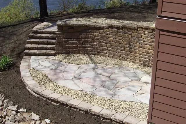 A stone patio with a curved retaining wall and a set of steps next to the side of a brown house.