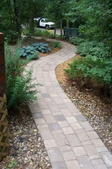 A winding walkway made of rectangular beige paving stones leads through a wooded area with gravel and lush greenery.