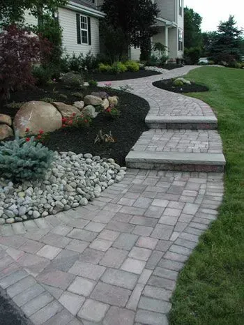 A winding paver walkway with two steps leads to a residential home entrance, flanked by garden beds with stones and shrubs.