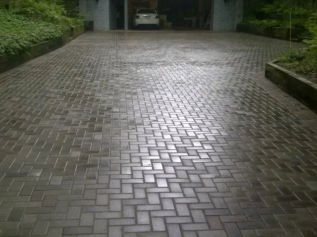 A wide, paved driveway with dark grey bricks laid in a herringbone pattern, leading toward a dark garage.