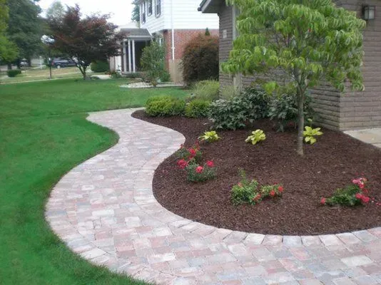 A curved brick walkway leads through a landscaped front yard with mulch beds, a small tree, and shrubs in front of a house.