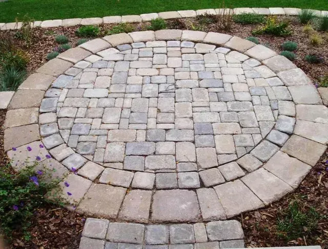 A circular stone paver patio sits in a garden bed with a border of lighter stone and a path leading to it.