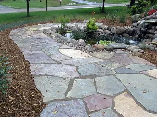A stone pathway curves beside a small, landscaped rock garden and water feature, surrounded by wood mulch.