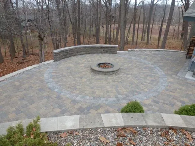 A stone patio with a circular fire pit, a curved stone seating wall, and a view of a wooded area in autumn.