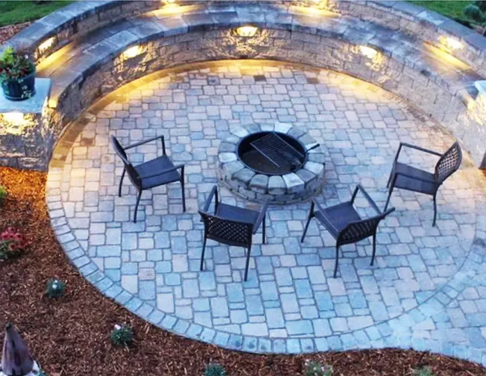 A circular stone patio features a central fire pit surrounded by four chairs and a curved, illuminated seating wall.