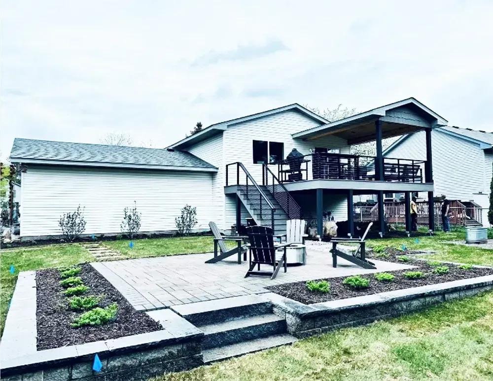 A backyard patio with four dark chairs surrounding a fire pit, set on pavers below a raised wooden deck with a staircase.