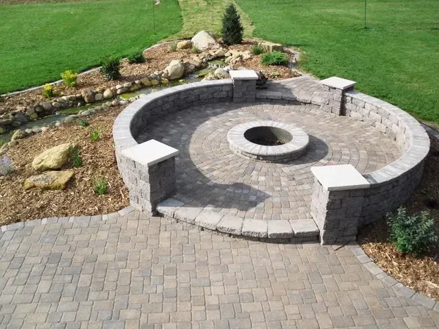 A circular paver patio with a central fire pit, bordered by a stone retaining wall with square pillars in a grassy yard.