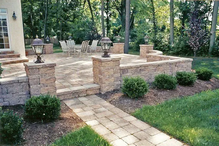 A stone patio with steps, seating, and outdoor lights, connected to a house by a stone walkway in a backyard.