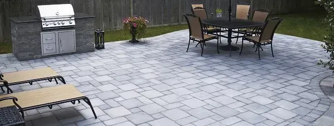 A stone patio featuring an outdoor kitchen, a dining table with chairs, and two lounge chairs on a patterned paver surface.