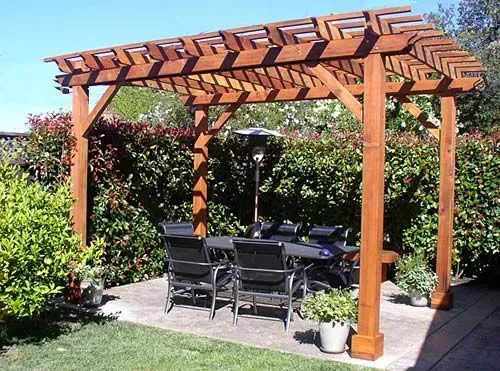 A wooden pergola stands on a stone patio, sheltering a dining table and chairs in a garden setting with hedges.