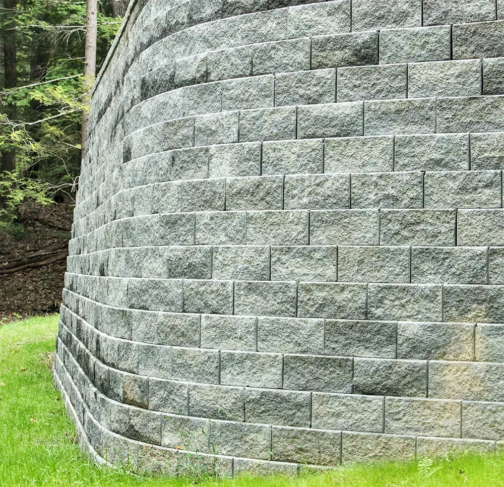 A curved retaining wall constructed of grey, textured concrete blocks, set against a background of green trees and grass.
