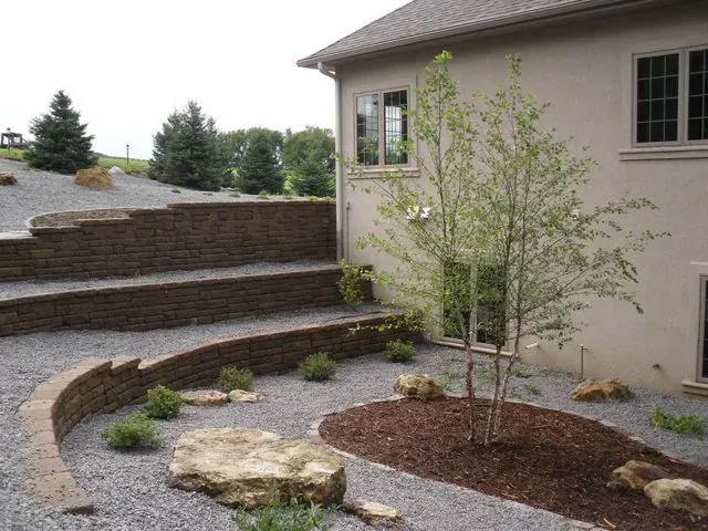 Tiered stone retaining walls, gravel beds, a birch tree, and landscape boulders outside a tan house.