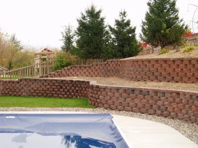 A two-tiered red stone retaining wall built into a sloped backyard behind a swimming pool.