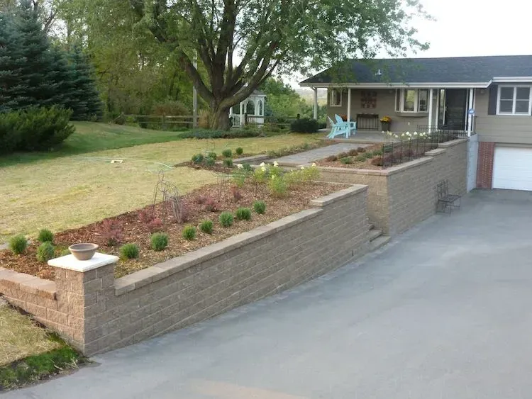A residential house featuring a two-tiered, light brown stone retaining wall bordering a driveway and front lawn.