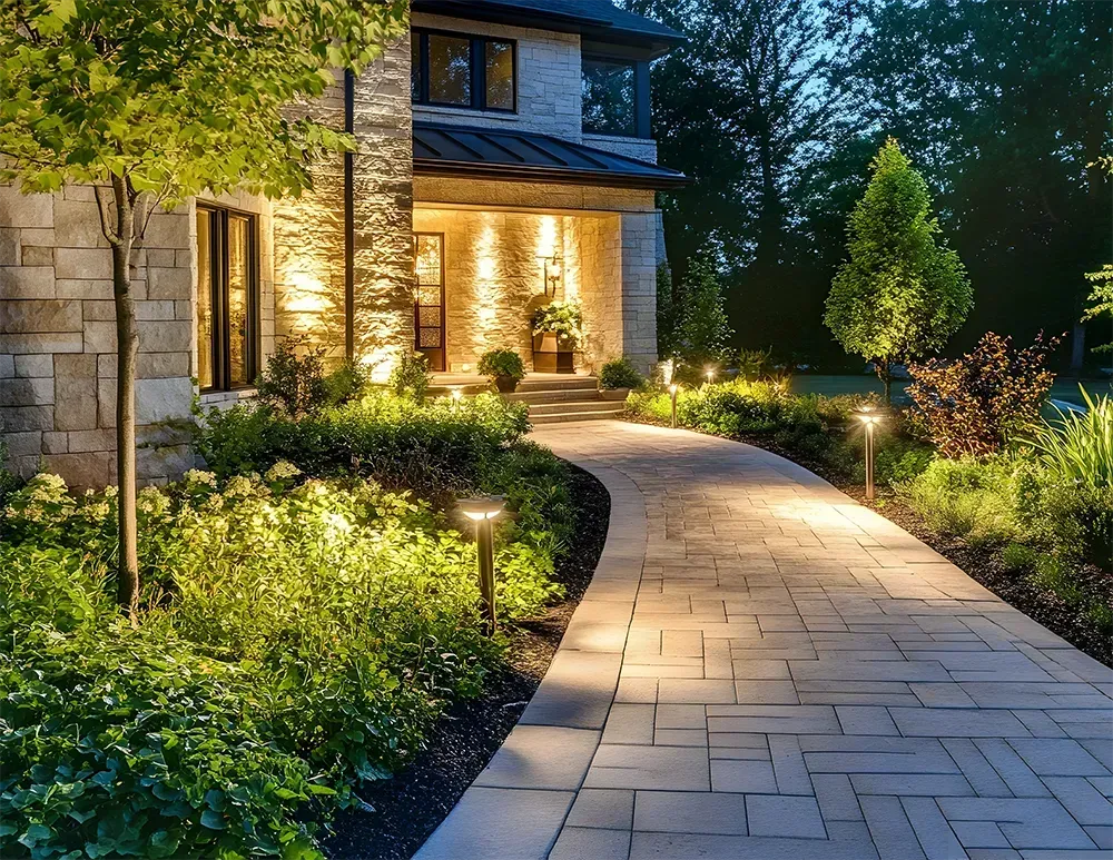 A stone walkway illuminated by low-profile lights leads to the entrance of a house at dusk, surrounded by landscaping.