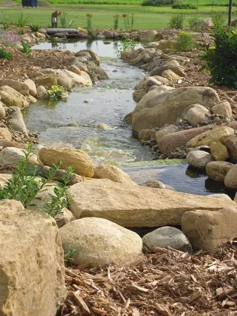 A man-made stone creek flows through a landscaped garden with wooden mulch and surrounding greenery.