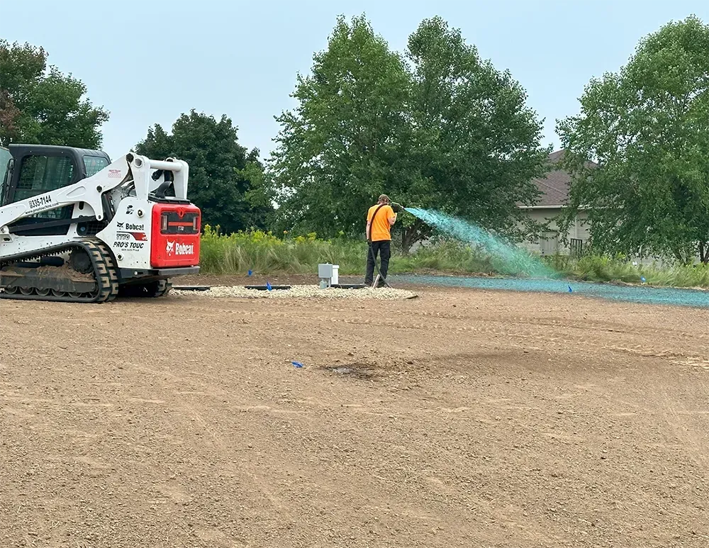 A worker in an orange shirt hydroseeding a dirt lot with a white Bobcat skid-steer nearby.