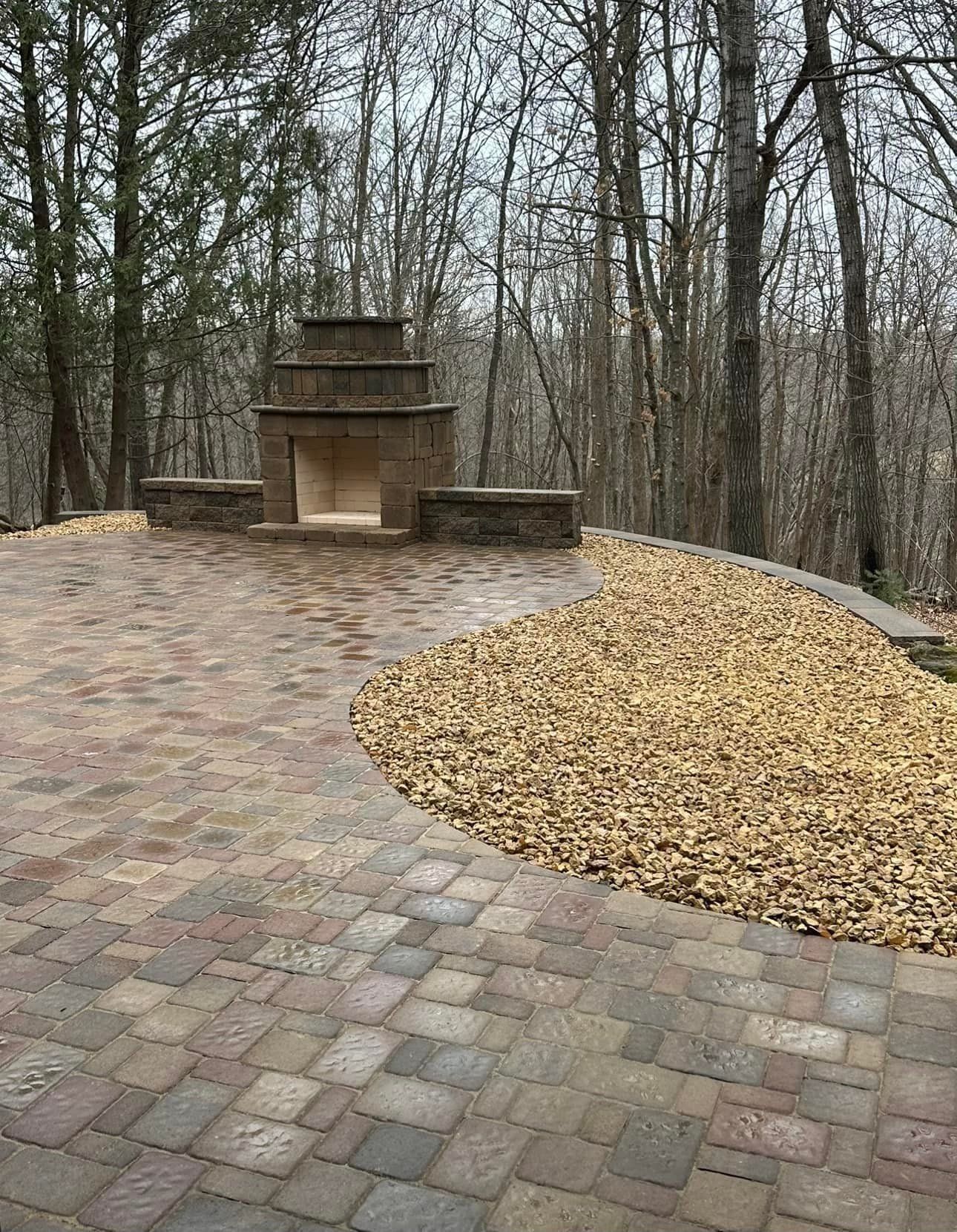 A tiered stone outdoor fireplace sits on a patio of mixed-tone pavers, bordered by a bed of beige gravel in a wooded area.