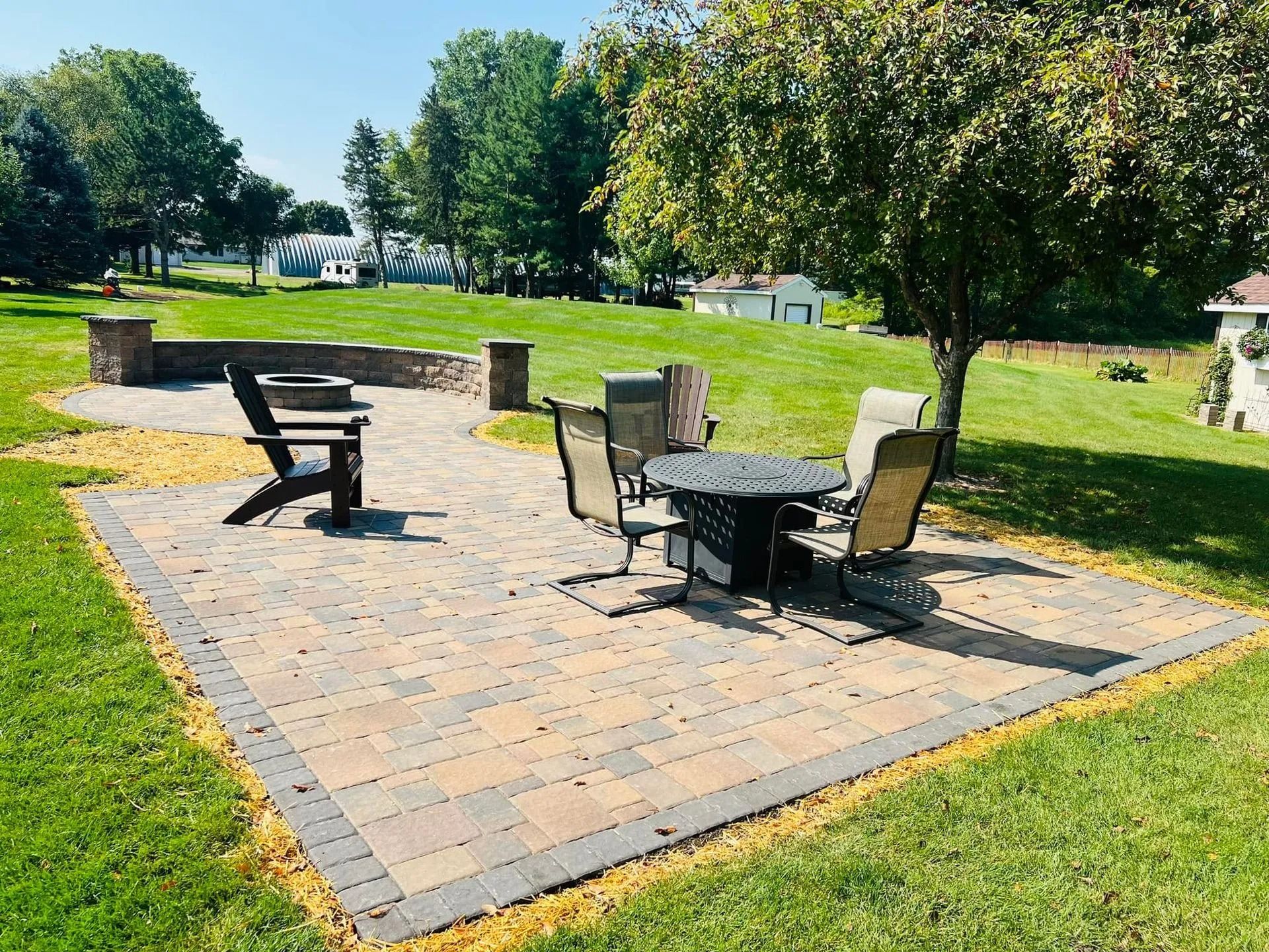A paved backyard patio with a fire pit area, a dining table with chairs, and surrounding grass on a sunny day.