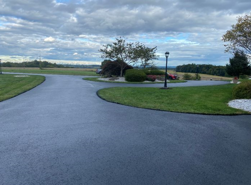 A black asphalt driveway surrounded by grass and trees on a cloudy day.
