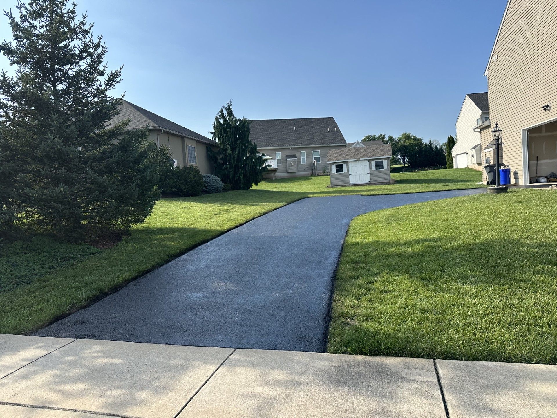 A freshly paved asphalt driveway leads toward a house, flanked by green grass, trees, and a small backyard shed.