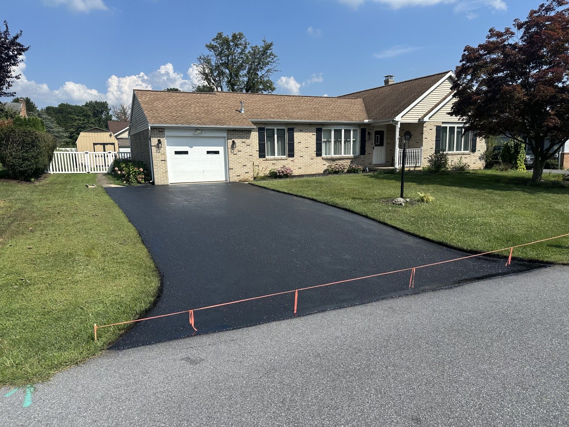 A freshly paved black asphalt driveway leads to a single-story brick house with a white garage door on a sunny day.