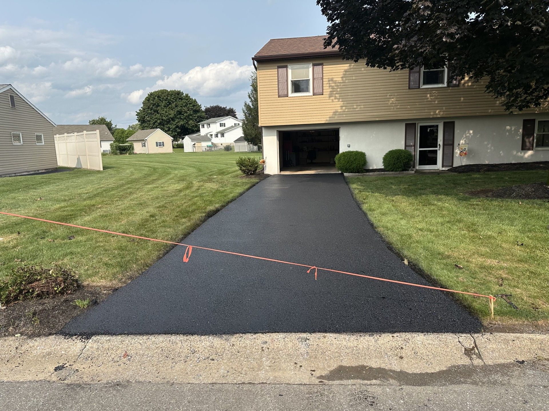 A newly paved asphalt driveway leads to the garage of a beige suburban home on a sunny day.