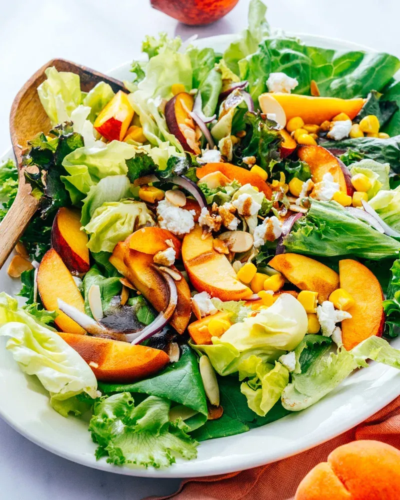 A salad with peaches , lettuce , corn , and almonds on a white plate with a wooden spoon.