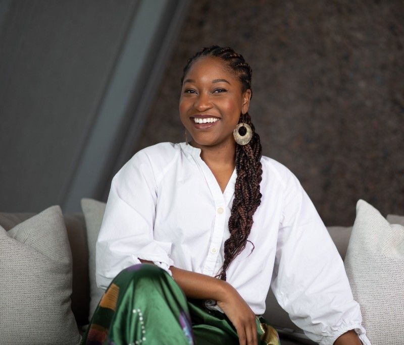 A woman is sitting on a couch wearing a white shirt and green pants.