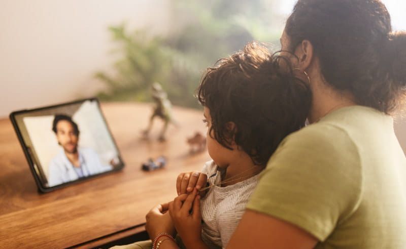 A woman is holding a child while having a video call with a doctor.