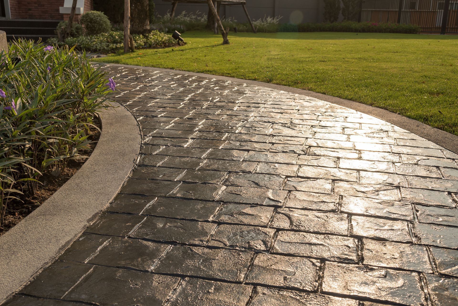 A dark, textured stone pathway curves through a lush green lawn, catching the sunlight.