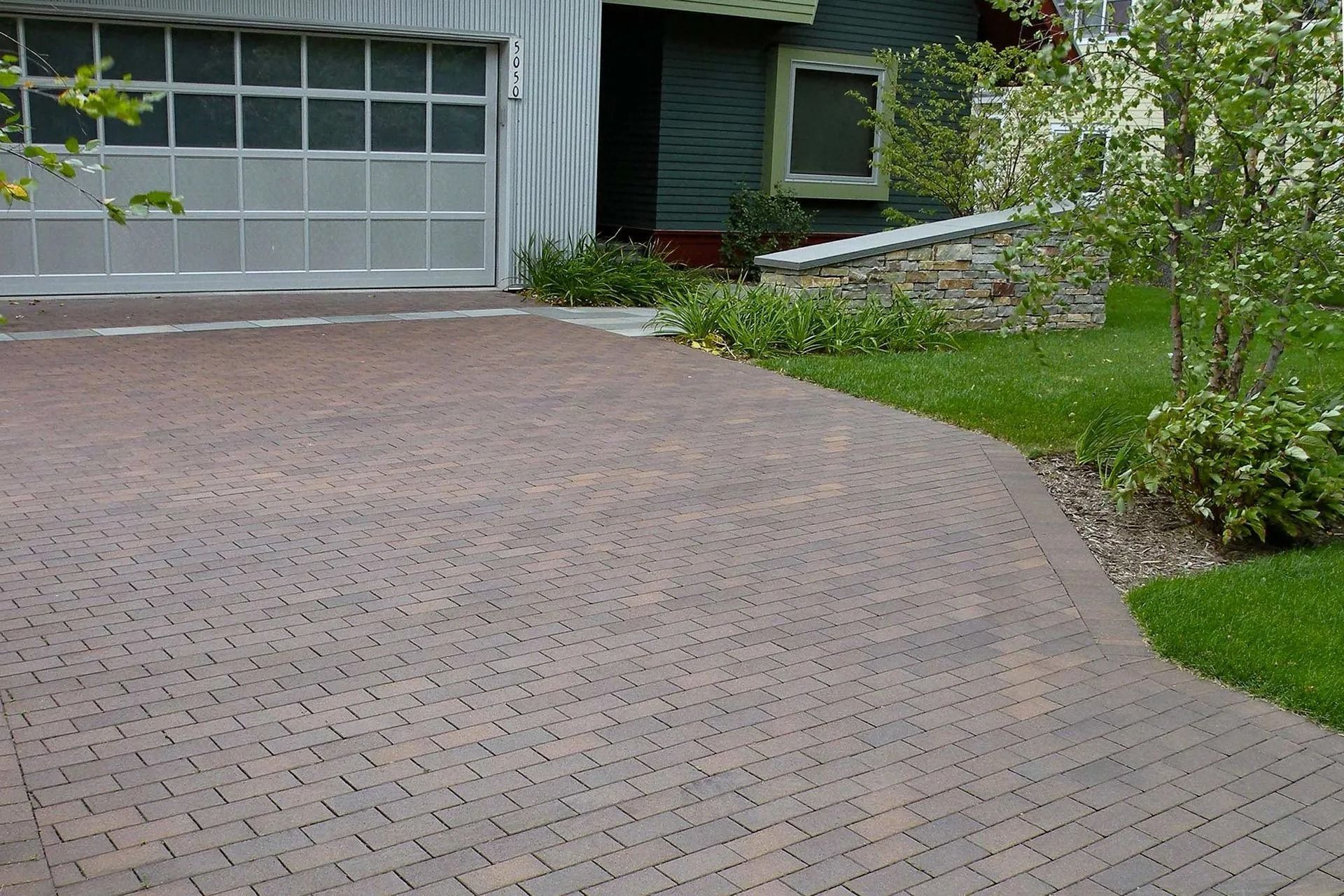 A brown brick driveway with a herringbone pattern leads to a modern home with a grey garage door and stone retaining wall.