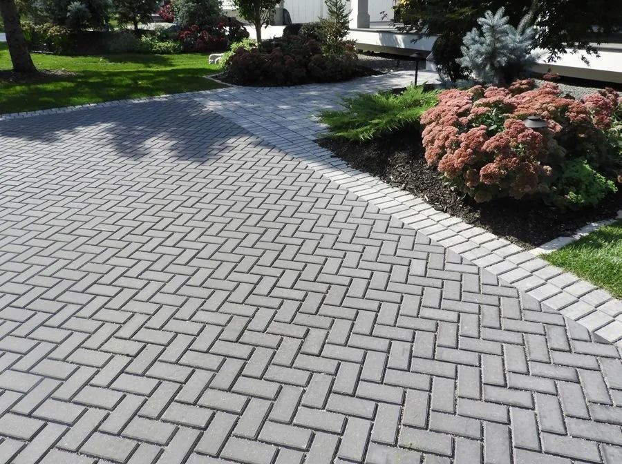 Gray herringbone-patterned stone pavers form a driveway lined with landscaped garden beds and green lawn.