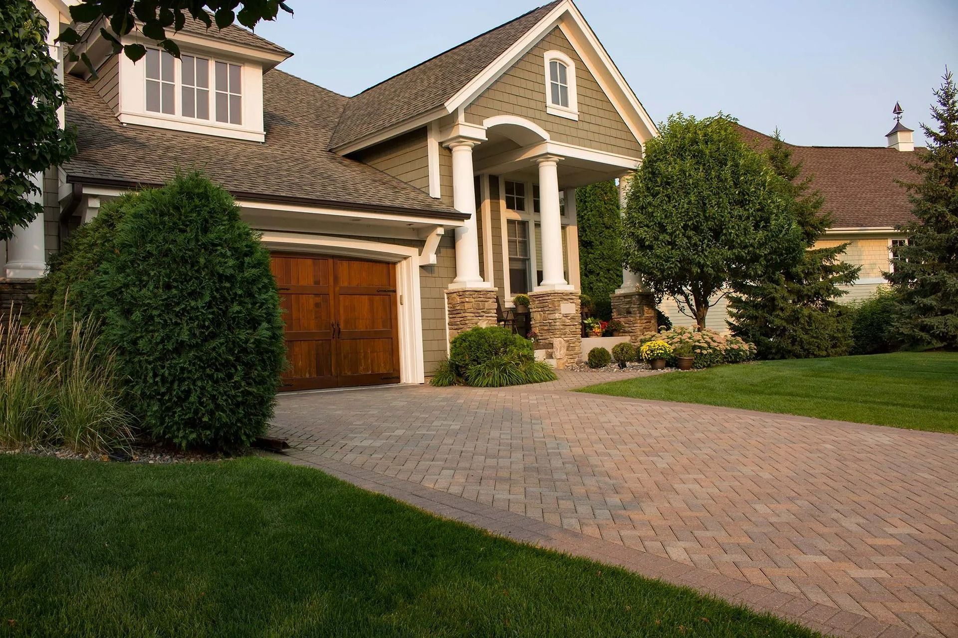 A house with a shingled roof, a wooden garage door, and a stone-accented porch, featuring a paved brick driveway.