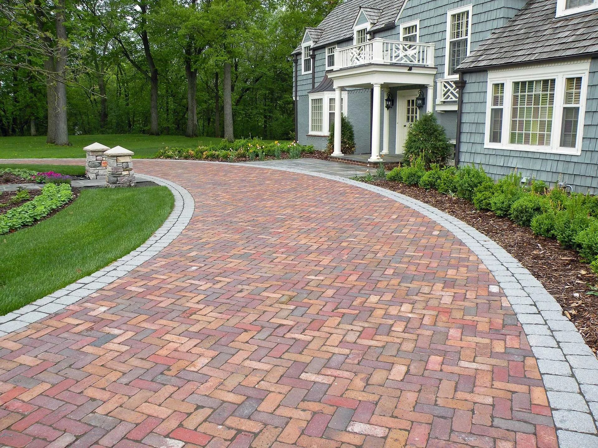 A curved brick driveway with a decorative gray stone border leads to the entrance of a blue house with lush green landscaping.