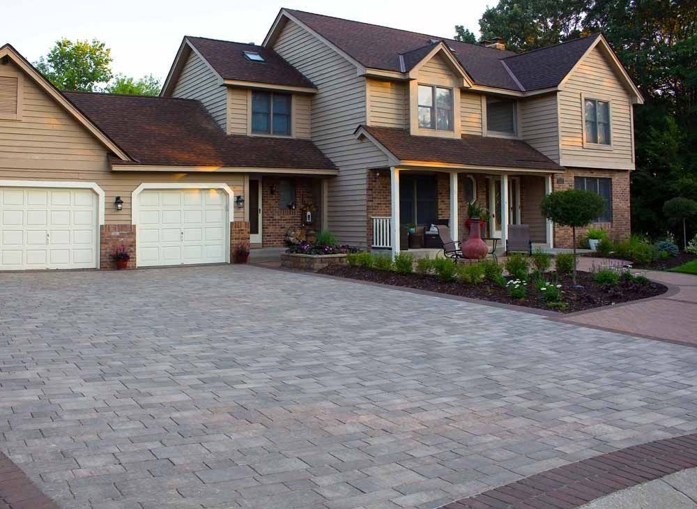 A large two-story suburban house with a beige facade, brown roof, and a patterned stone-paver driveway and front patio.