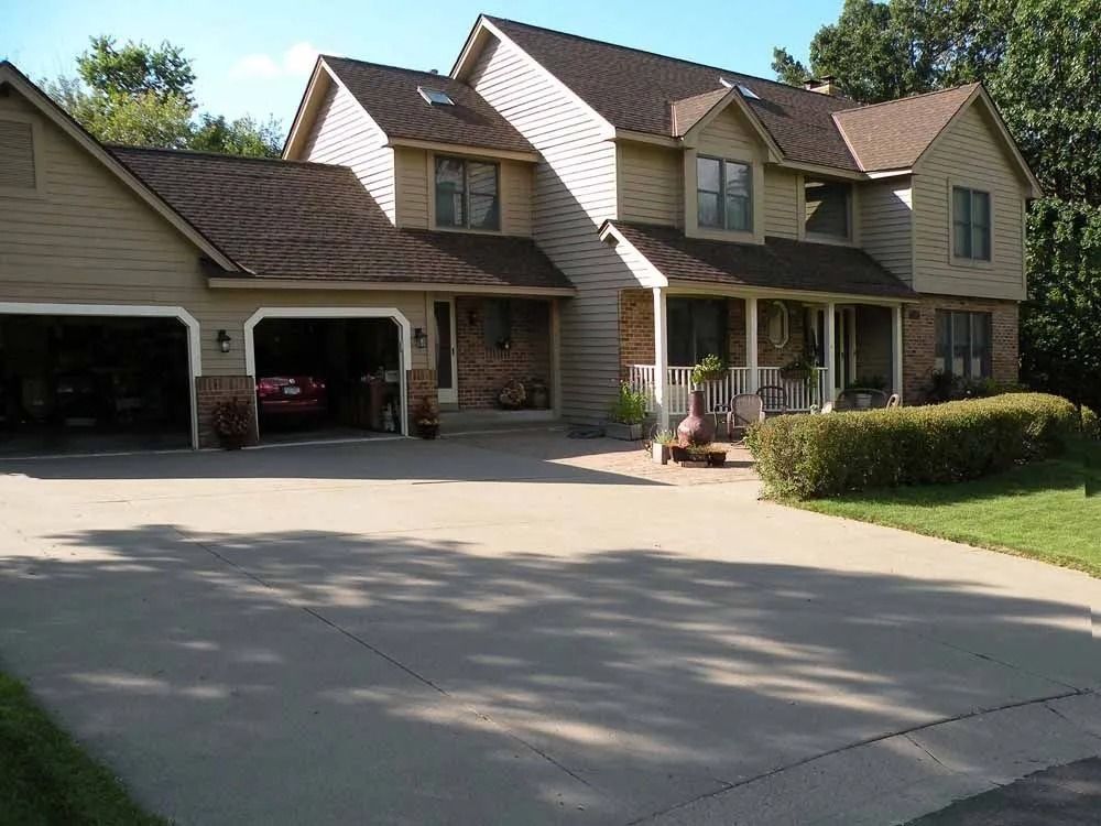 A two-story tan house with a dark shingled roof, a front porch, and a two-car garage at the end of a concrete driveway.