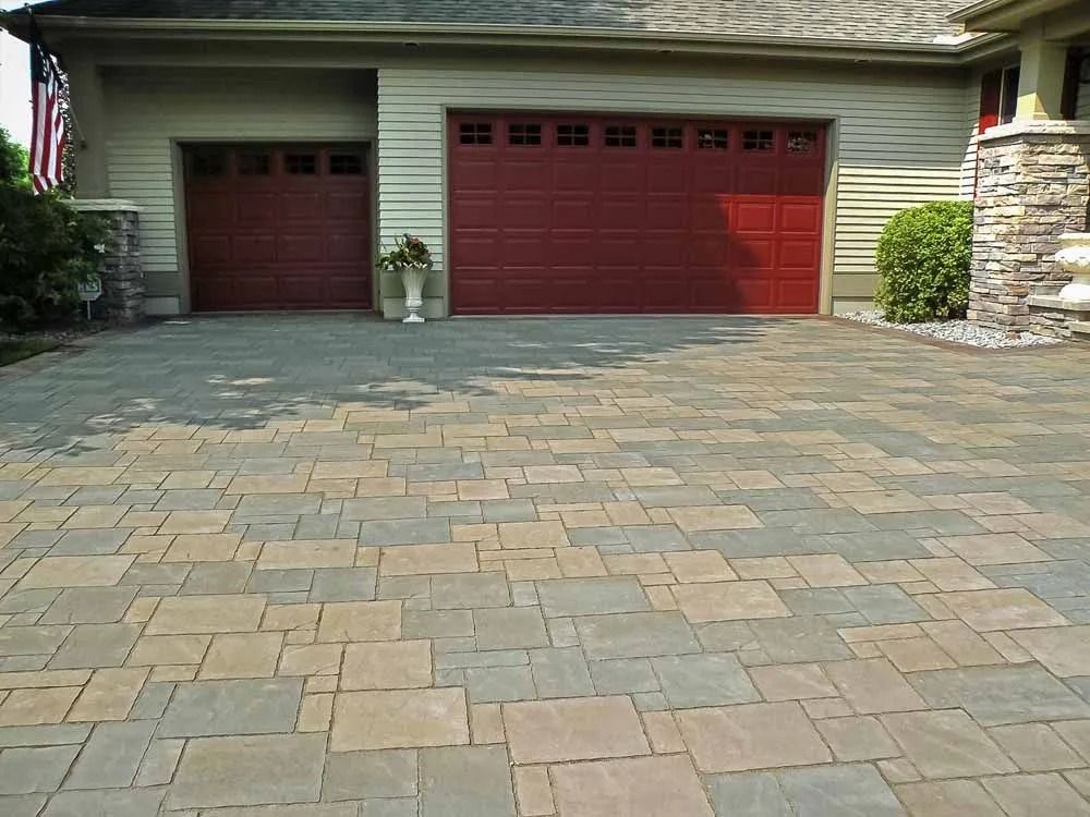A multi-tonal paver driveway leads to a house with two dark red garage doors and light tan siding.