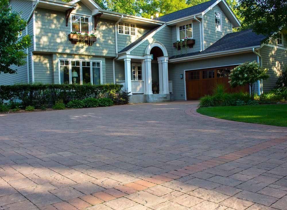 A suburban home with a green exterior, arched entryway, and a large, paved brick driveway on a sunny day.
