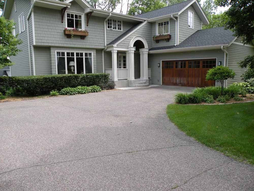 A gray two-story house with a wooden garage door and a large paved driveway surrounded by landscaping.