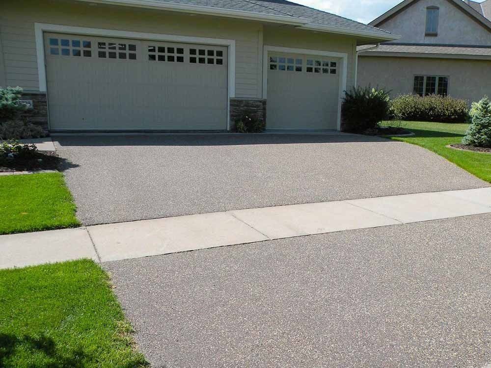 A wide, beige suburban house with two garage doors, a textured driveway, and a sidewalk in the foreground.