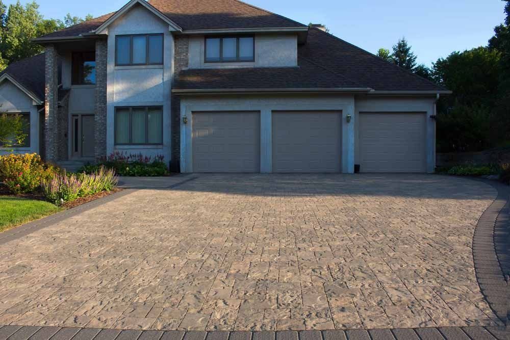 A two-story suburban home featuring a beige stone-paved driveway, a three-car garage, and light gray exterior siding.