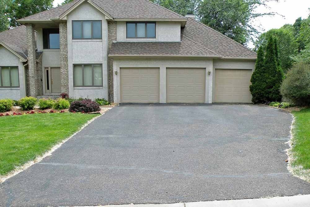 A suburban two-story house with a three-car garage and a wide asphalt driveway surrounded by a green lawn.