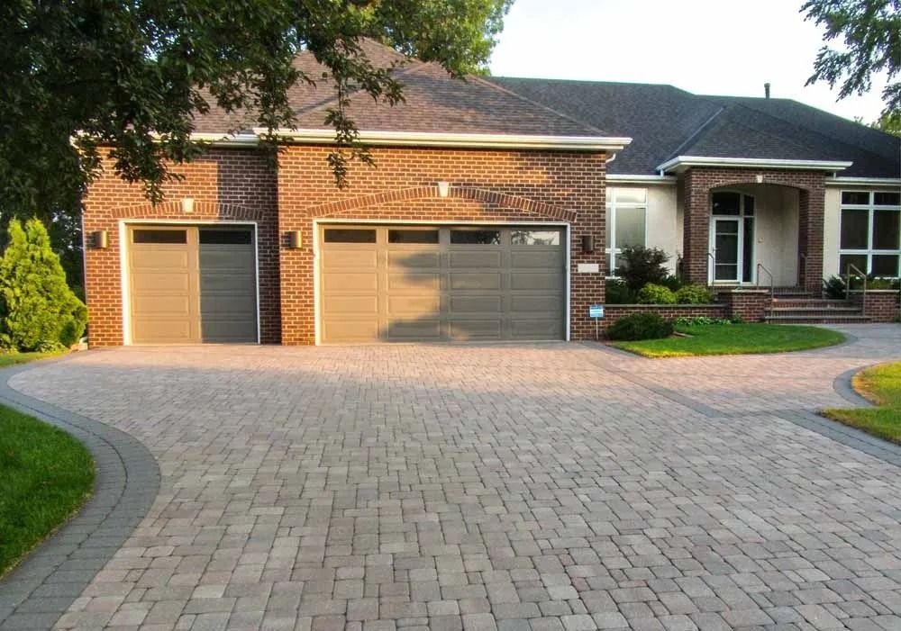 A brick house with a paved driveway, two garage doors, and a small front entryway, set against a backdrop of trees.
