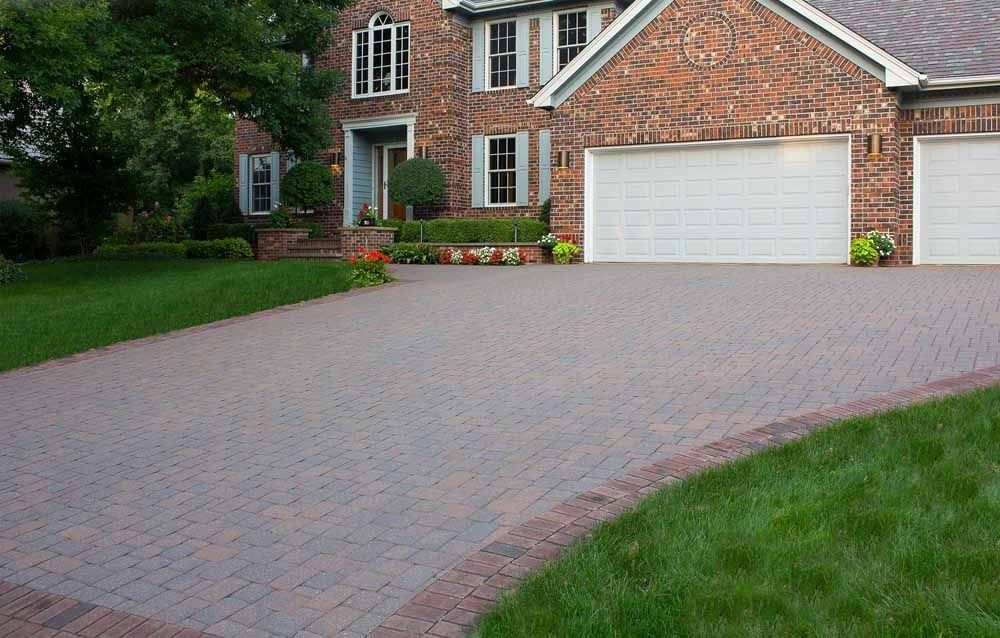 A brick paved driveway leads to a brick house with white garage doors and green lawn areas on a sunny day.