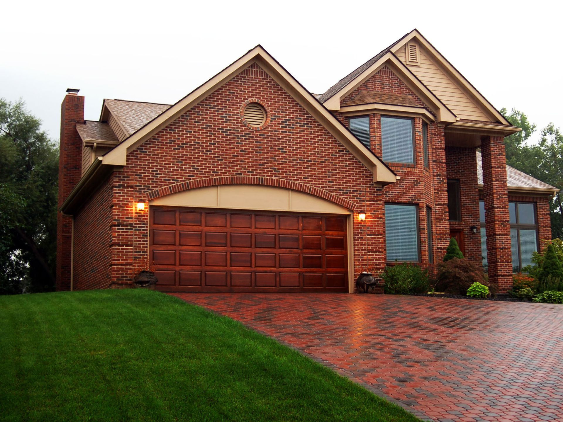 A two-story brick house with a large wooden garage door, a paved circular driveway, and a manicured green lawn.