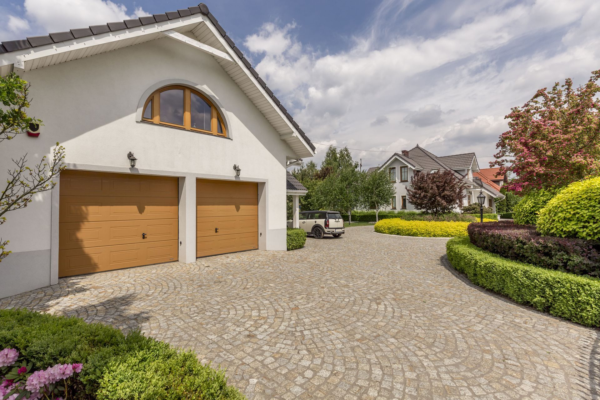 A house with two wood-paneled garage doors and an arched window, featuring a cobblestone driveway and landscaped garden.